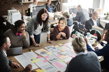 Group of people at table in office.  Table is covered in printouts with sticky notes.  One person is standing up with their hands on the table leading the discussion.